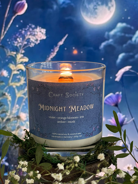 A scented candle called Midnight Meadow  on a floral and night background, lit, clear glass jar, deluxe version with wooden wick