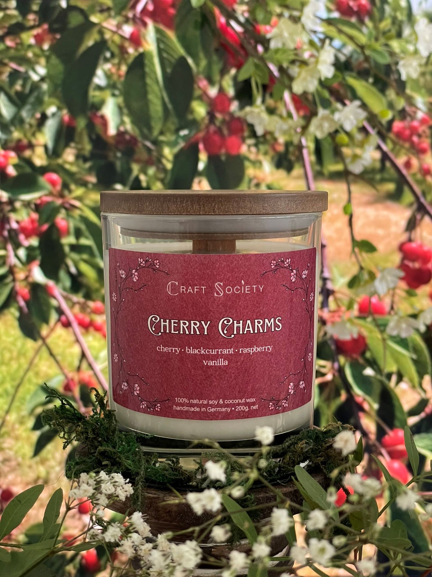 A scented candle called Cherry Charms on a floral and nature background, boxed, clear glass jar, deluxe version with wooden wick