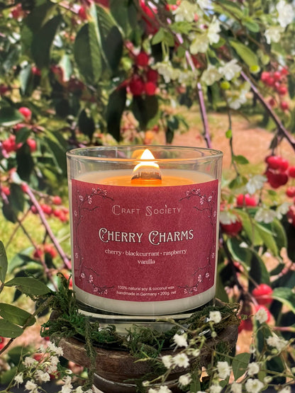 A scented candle called Cherry Charms on a floral and nature background, lit, clear glass jar, deluxe version with wooden wick
