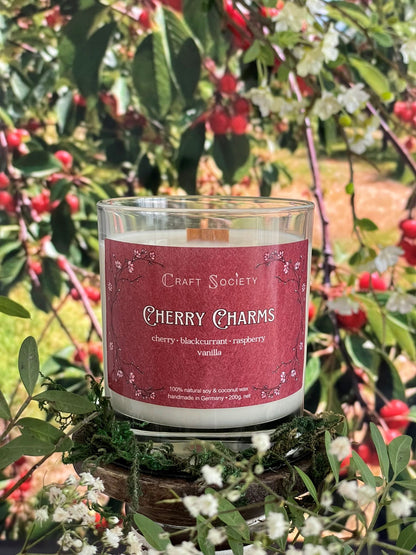 A scented candle called Cherry Charms on a floral and nature background, unlit, clear glass jar, deluxe version with wooden wick
