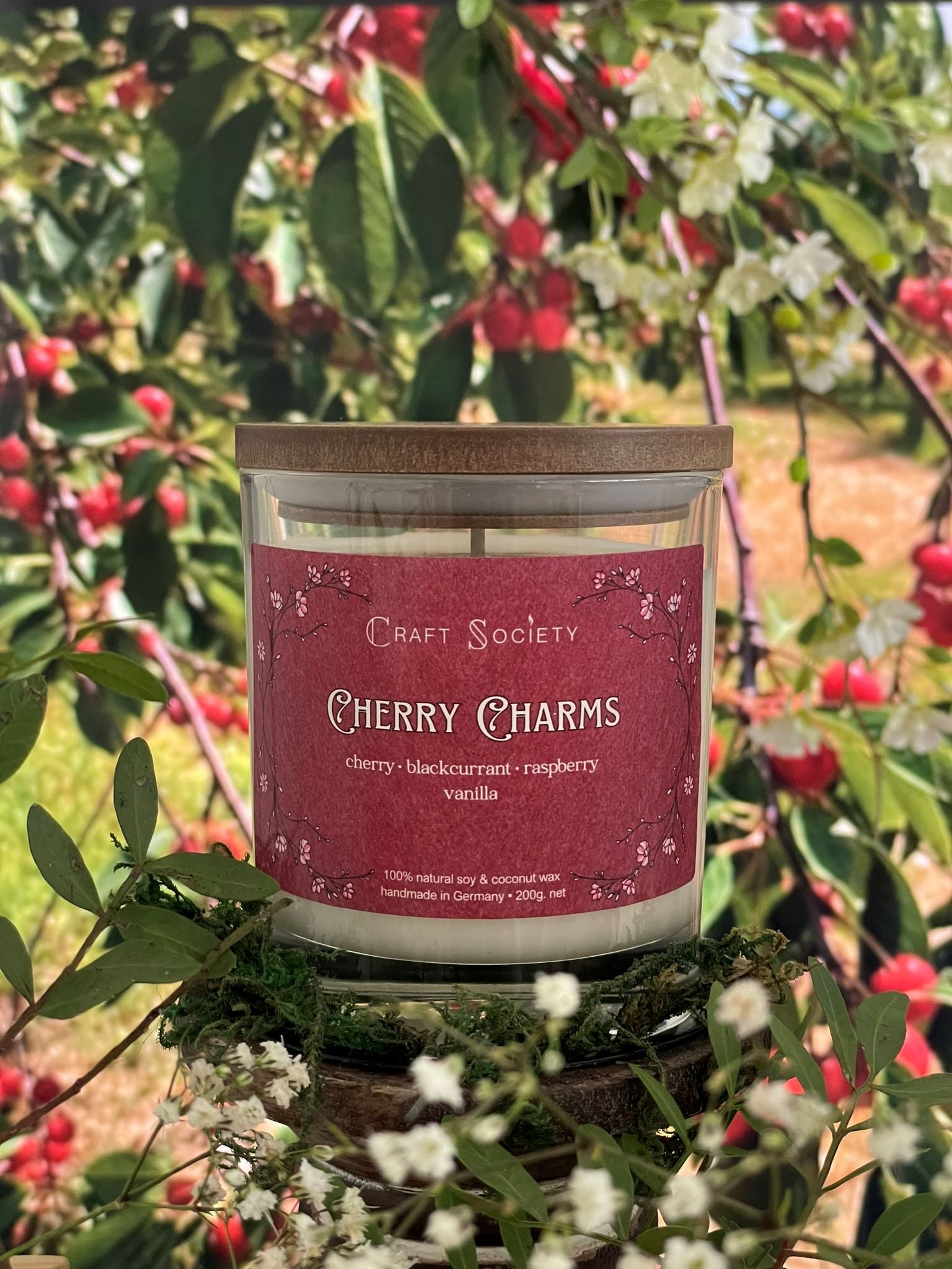 A scented candle called Cherry Charms on a floral and nature background, boxed, clear glass jar, regular version with cotton wick