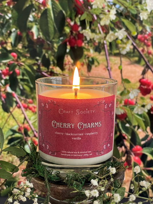 A scented candle called Cherry Charms on a floral and nature background, lit, clear glass jar, regular version with cotton wick