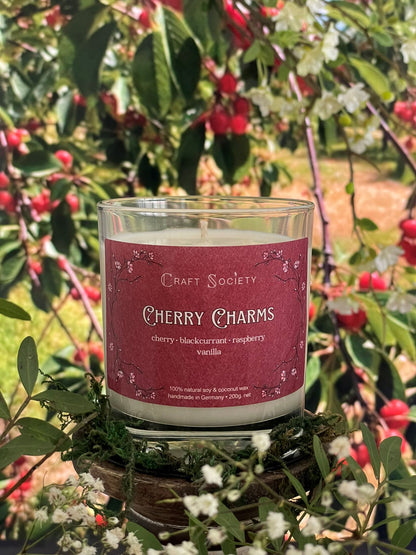 A scented candle called Cherry Charms on a floral and nature background, unlit, clear glass jar, regular version with cotton wick