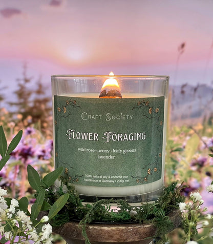 A scented candle called Flower Foraging on a floral and sunrise background, lit, clear glass jar, deluxe version with wooden wick