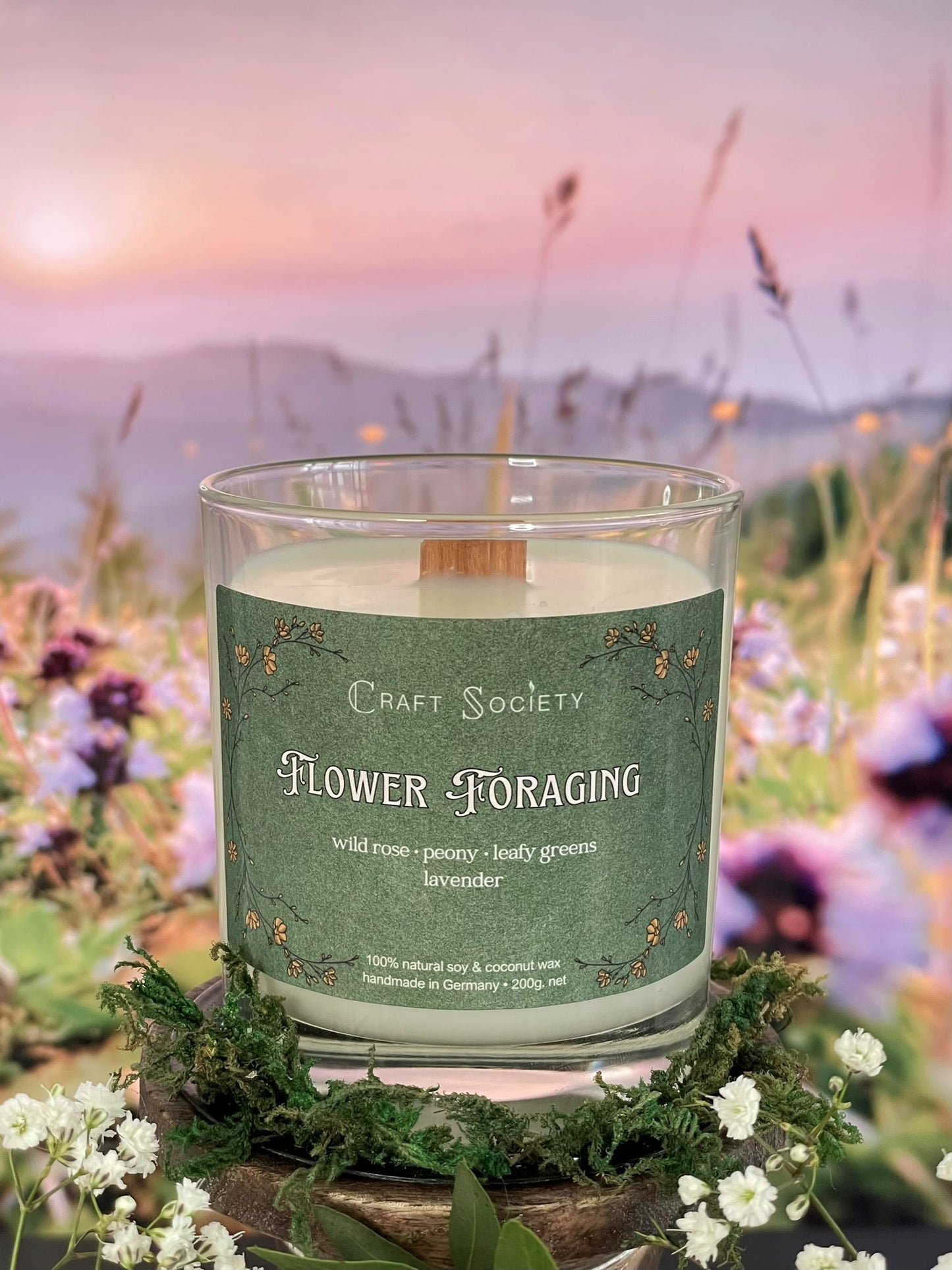 A scented candle called Flower Foraging on a floral and sunrise background, unlit, clear glass jar, deluxe version with wooden wick
