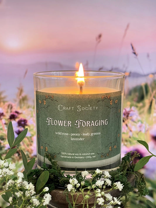 A scented candle called Flower Foraging on a floral and sunrise background, lit, clear glass jar, regular version with cotton wick