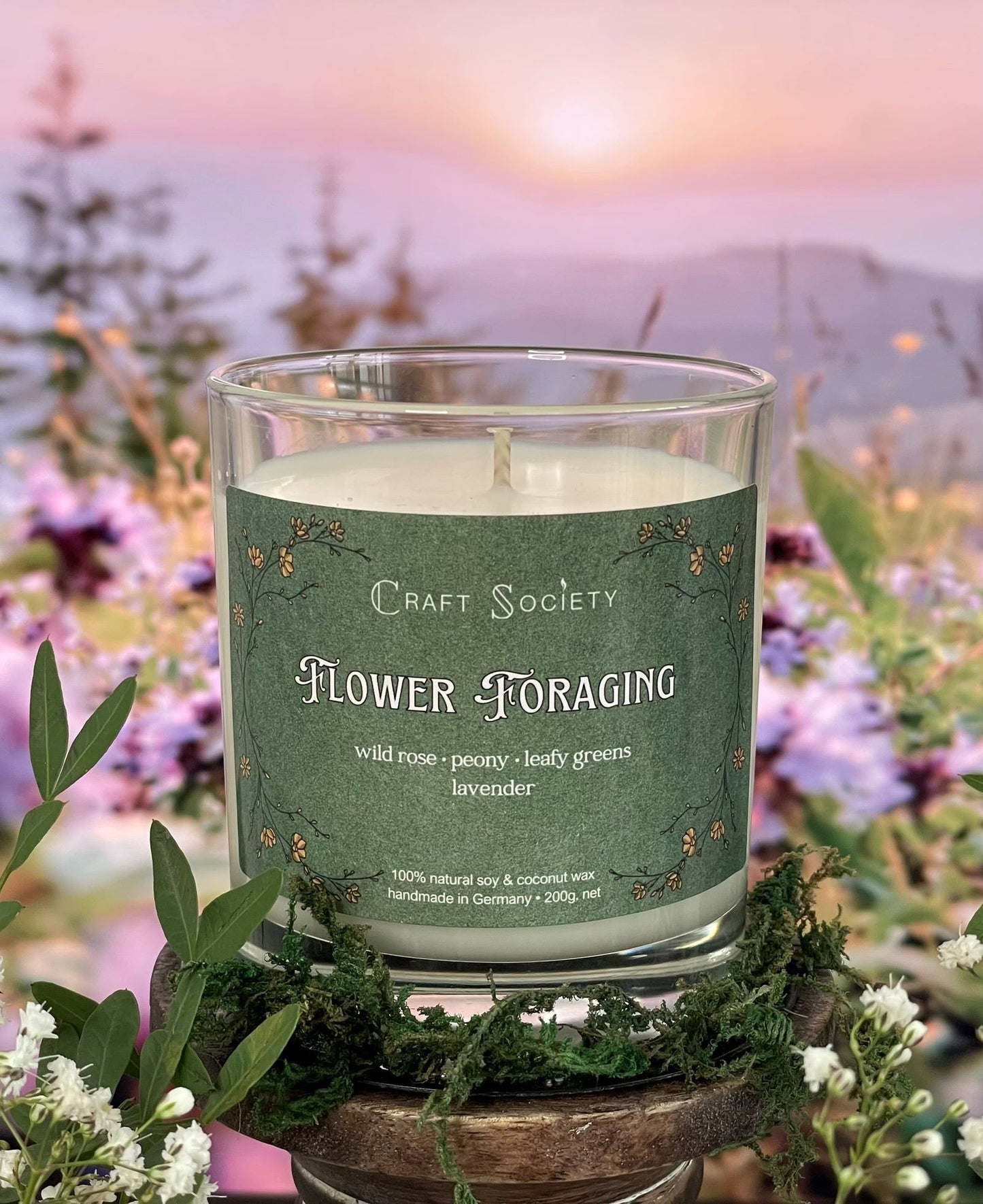 A scented candle called Flower Foraging on a floral and sunrise background, unlit, clear glass jar, regular version with cotton wick