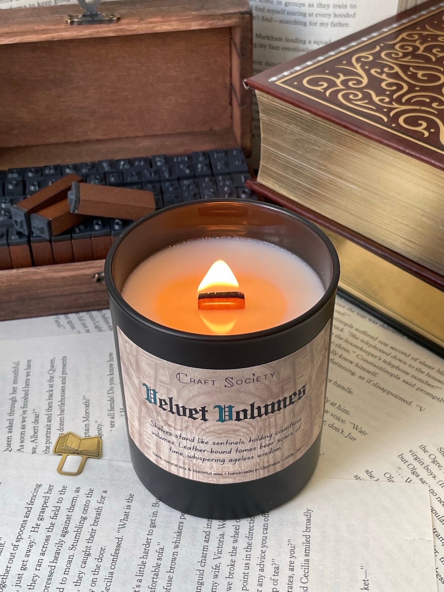 A burning deluxe scented candle in a black jar on a decorated background with books flowers and texts