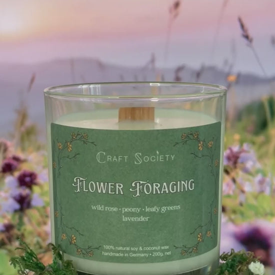 A scented candle called Flower Foraging on a floral and sunrise background, being lit, clear glass jar, deluxe version with wooden wick