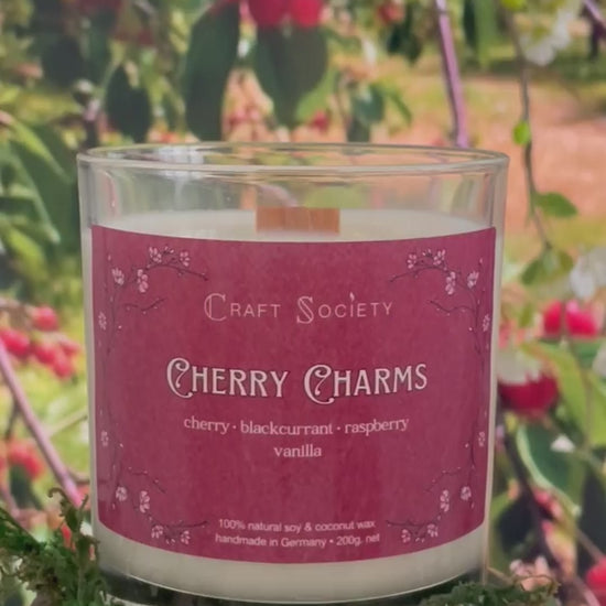 A scented candle called Cherry Charms on a floral and nature background, being lit, clear glass jar, deluxe version with wooden wick