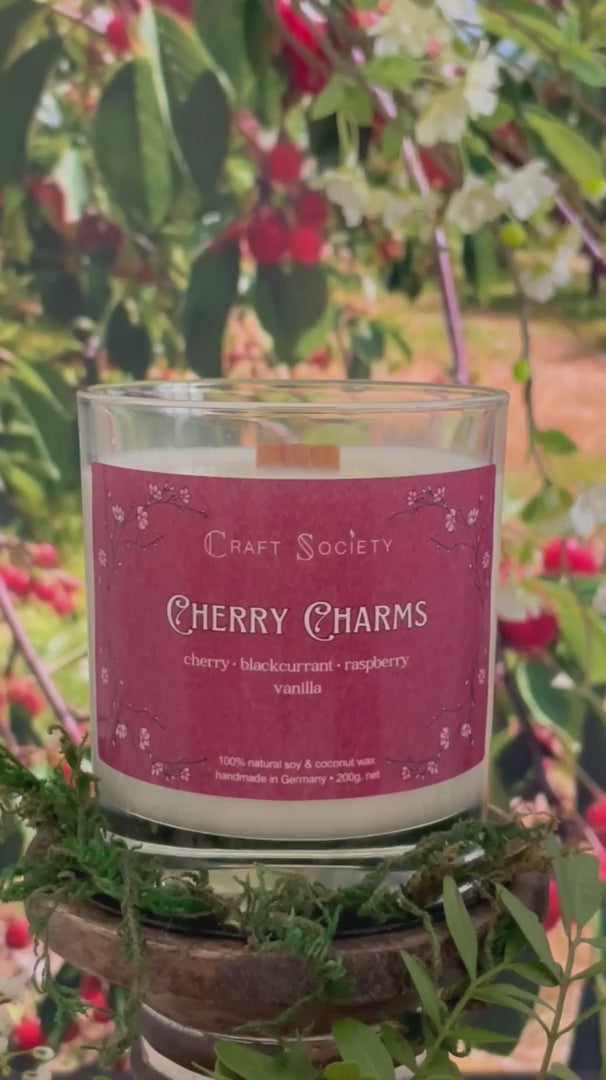 A scented candle called Cherry Charms on a floral and nature background, being lit, clear glass jar, deluxe version with wooden wick