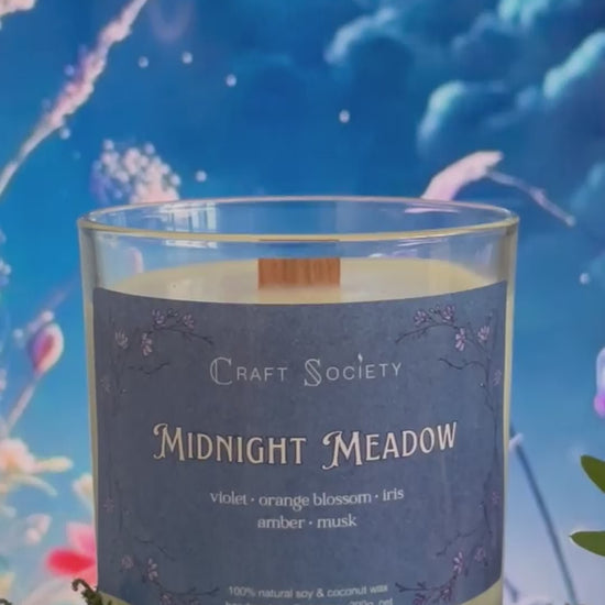 A scented candle called Midnight Meadow on a floral and night background, being lit, clear glass jar, deluxe version with wooden wick