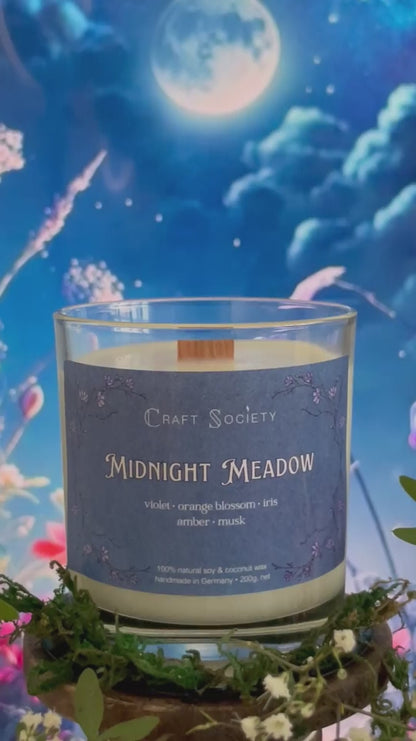 A scented candle called Midnight Meadow on a floral and night background, being lit, clear glass jar, deluxe version with wooden wick