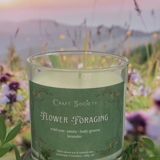 A scented candle called Flower Foraging on a floral and sunrise background, being lit, clear glass jar, regular version with cotton wick
