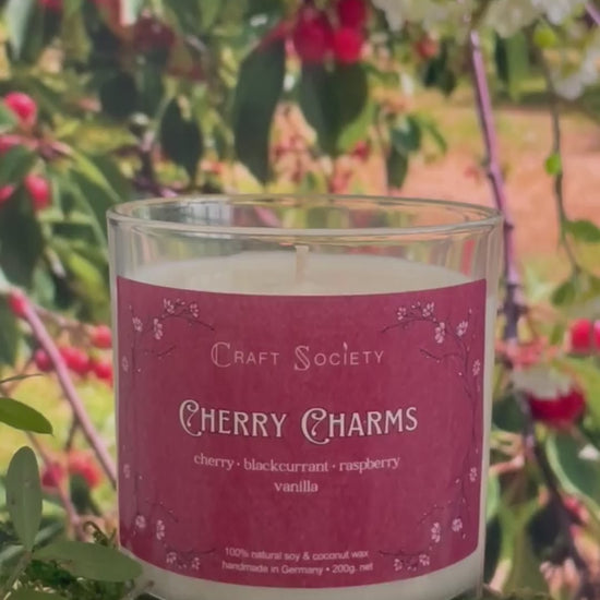 A scented candle called Cherry Charms on a floral and nature background, being lit, clear glass jar, regular version with cotton wick