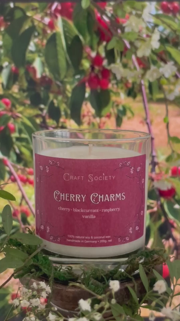 A scented candle called Cherry Charms on a floral and nature background, being lit, clear glass jar, regular version with cotton wick